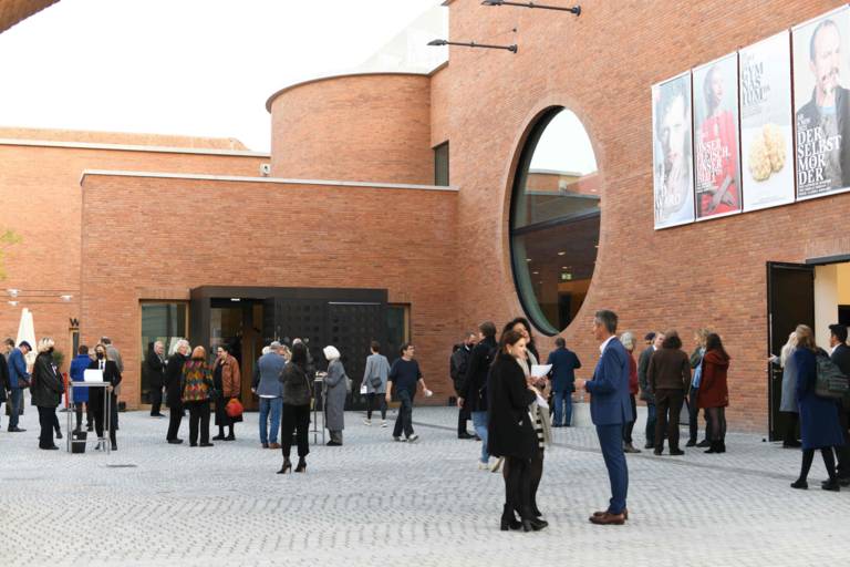 Theatergäste halten sich vor der Vorstellung im Innenhof des Volkstheaters in München mit Backsteinfassade auf.